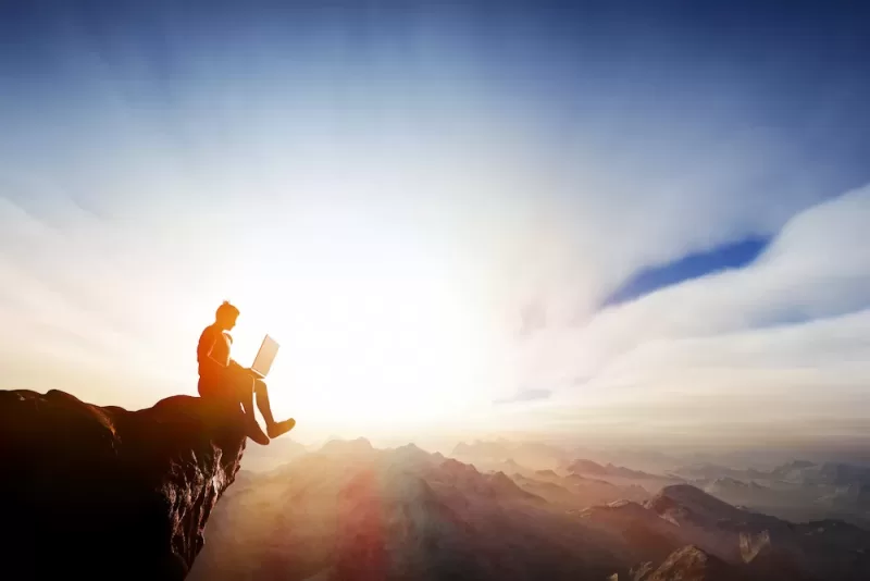 An adventure-seeking student utilizes a laptop while sitting at a cliff's edge, emphasizing the flexibility and possibilities of online learning.